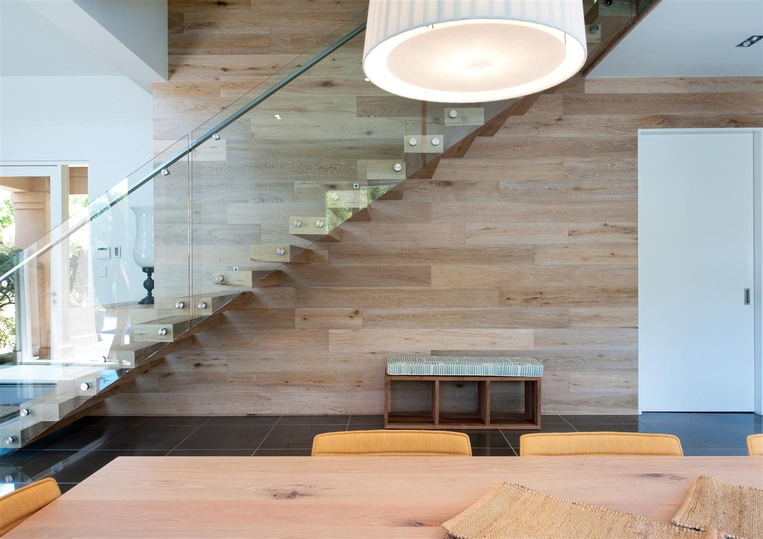 Interior — a floating timber stair beside the dining area, beneath a large circular pendant, with a glass balustrade